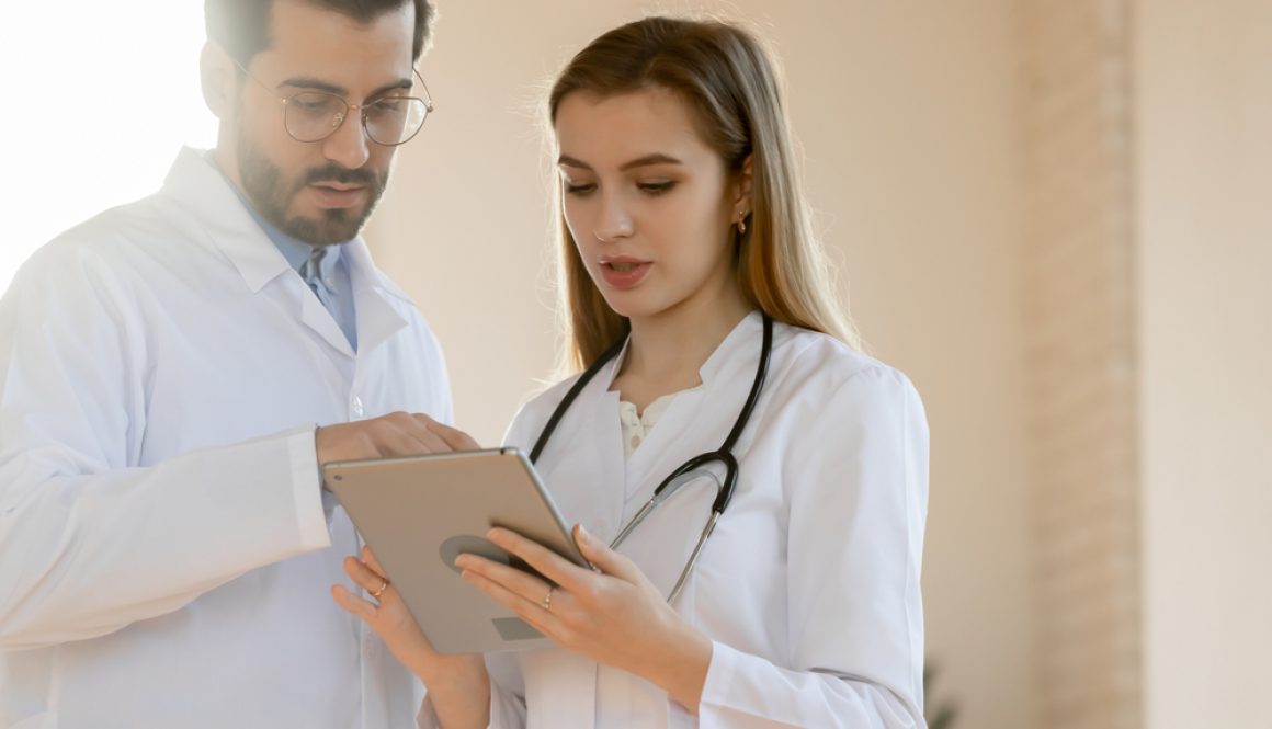 Two,Diverse,Doctors,In,White,Medical,Uniforms,Look,At,Tablet